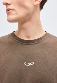 Νέος άνθρωπος που φοράει καφέ μπλούζα με λαιμόκοψη και μικρό λευκό αφηρημένο λογότυπο στο κέντρο του στήθους, κοιτάζοντας αριστερά μπροστά σε απλό φόντο.