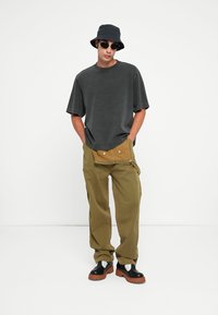 T-shirt gris surdimensionné, pantalon en velours côtelé vert olive avec poches avant, chapeau seau noir, lunettes de soleil, et chaussures à plateforme noires avec semelles brunes.