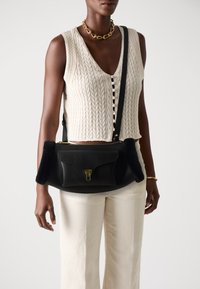 Borsa a tracolla in pelle nera con finitura strutturata, con morbidi accenti laterali neri e una chiusura dorata. Indossata con un top in maglia beige e pantaloni chiari.