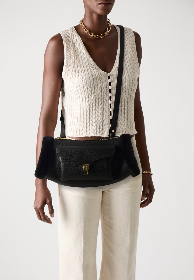 Borsa a tracolla in pelle nera con finitura strutturata, con morbidi accenti laterali neri e una chiusura dorata. Indossata con un top in maglia beige e pantaloni chiari.