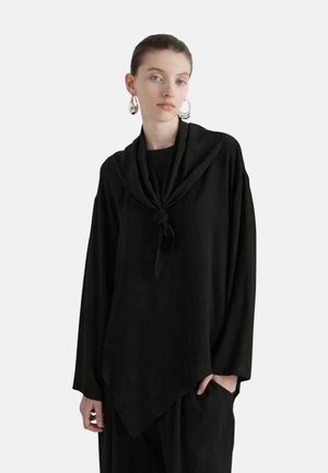 Jeune femme aux cheveux plaqués en arrière porte un haut noir oversize à manches longues avec un détail de foulard noué et de grandes boucles d'oreilles créoles argentées.