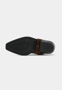 Μαύρη λαστιχένια σόλα με μυτερή μύτη και λεπτομέρεια με καφέ δερμάτινο λουράκι σε σόλα επίσημου υποδήματος, σε θέα από κάτω σε λευκό φόντο.