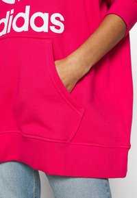 Růžový oversized svetr s bílým logem, který má přední kapsu, měkkou texturu a volný střih, spárovaný se světle modrými džíny.