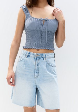 Mujer con un top corto azul fruncido con detalle de lazo y shorts de mezclilla claros de pierna ancha y talle alto, de pie frente a un fondo liso.