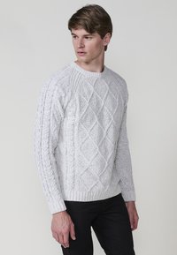 Witte kabelgebreide sweater met een ronde halslijn, getextureerde ruitpatronen en geribde manchetten. Draagt met zwarte broek.
