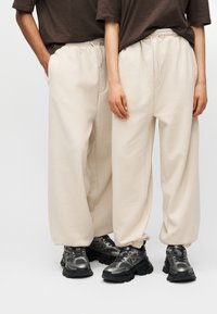 Μπεζ φόρμες με ελαστική μέση και πλαϊνές τσέπες, συνδυασμένες με μαύρα αθλητικά παπούτσια με χοντρή σόλα και ασημί λεπτομέρειες.