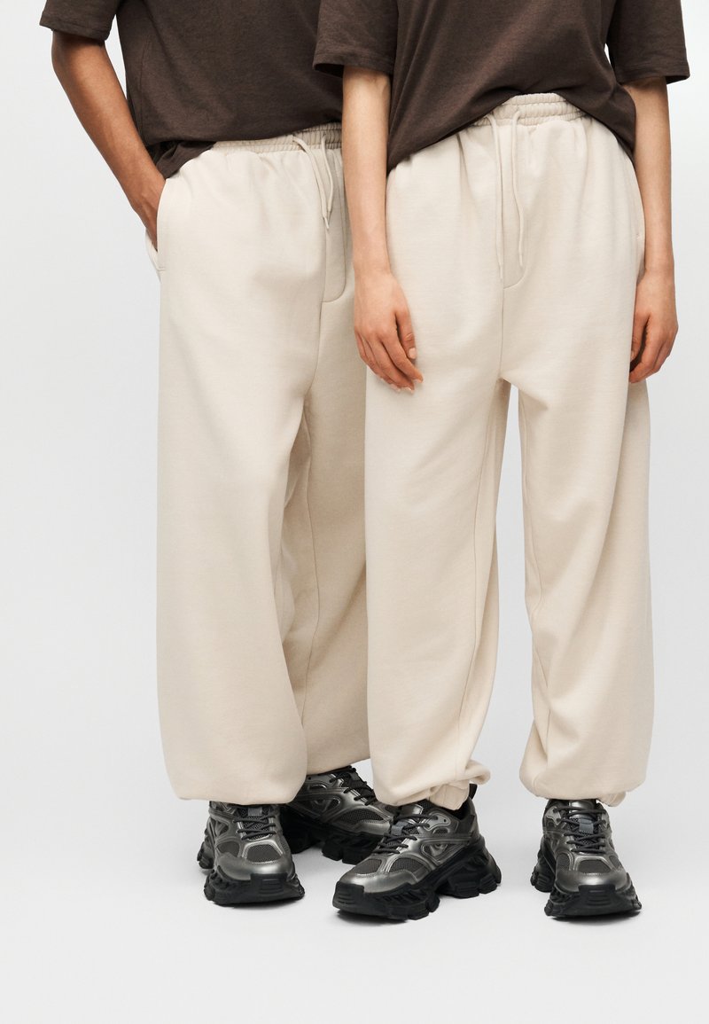 Μπεζ φόρμες με ελαστική μέση και πλαϊνές τσέπες, συνδυασμένες με μαύρα αθλητικά παπούτσια με χοντρή σόλα και ασημί λεπτομέρειες.