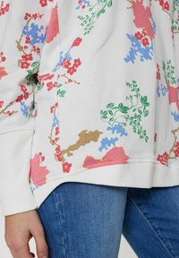 Vit sweatshirt med ett blommönster i rött, rosa, blått och grönt. Ribbat manschetter och nederkant, med en rundad bottenkant. Matchas med blå jeans.