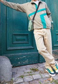 Beige fleece pullover med teal accenter, lyse beige bukser og multicolor sneakers, parret med en lille grøn crossbody taske.