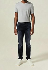 Homme portant un t-shirt gris clair à manches courtes, un jean slim foncé avec des revers et des baskets blanches, debout devant un fond uni.
