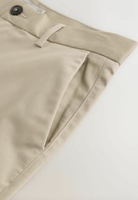 Pantalones beige doblados mostrando la pretina con botón negro, trabilla para cinturón y bolsillo lateral sobre un fondo claro.