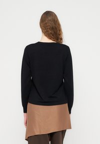 Μαύρο πουλόβερ με μακριά μανίκια, ριμπ μανσέτες και στρογγυλή λαιμόκοψη, φορεμένο πάνω από καφέ φούστα με ασύμμετρο τελείωμα. Λεία υφή πλέξης.