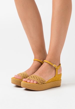 Sandales à talon compensé jaune avec un empeigne tissée, une bride de cheville réglable, une semelle intermédiaire texturée et un talon fermé. Semelle plate avec des bords patternisés.