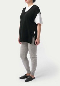 Svart stickad ärmlös tröja med V-ringning, sidoflikar och rosettdetaljer, bärs över en vit T-shirt och matchas med grå leggings och svarta tofflor.