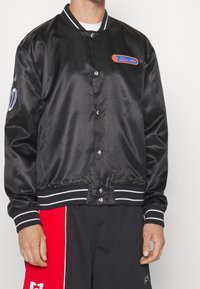 Black satin bomber jacket with snap buttons, striped cuffs and hem, featuring a blue and orange logo patch on the left chest.