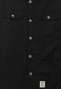 Czarna koszula zapinana na guziki, z dwoma klapami na kieszeniach na piersi oraz małą białą metką z logo przedstawiającym sylwetkę łosia i nazwą marki, umieszczoną u dołu.