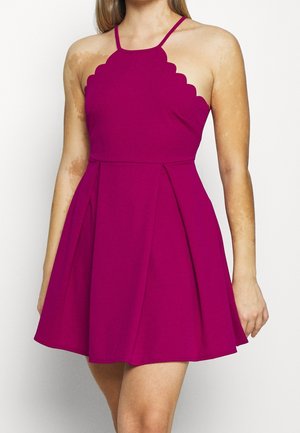 Femme portant une robe magenta sans manches avec un col festonné et une jupe plissée en ligne A, debout devant un fond blanc uni.