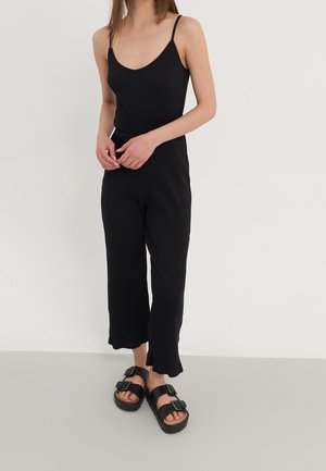 Vrouw in een mouwloos zwart geribbeld topje en bijpassende wijde broek met zwarte buckle sandalen, staand tegen een effen achtergrond.