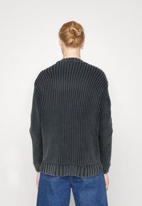 Pull en maille épaisse gris foncé avec texture côtelée, épaules tombantes et col rond, associé à un jean en denim bleu.