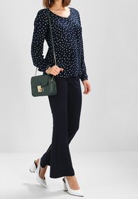 Blouse marine avec un motif floral blanc, manches longues ; associée à un pantalon noir évasé et des chaussures à talons blocs blanches ; portant un sac vert.