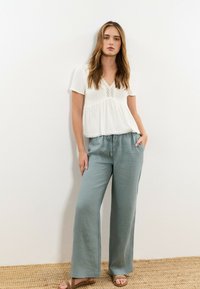 Mujer joven con cabello suelto y castaño, viste una blusa blanca de manga corta, pantalones anchos azul claro con cordón y sandalias marrones, de pie frente a una pared blanca.