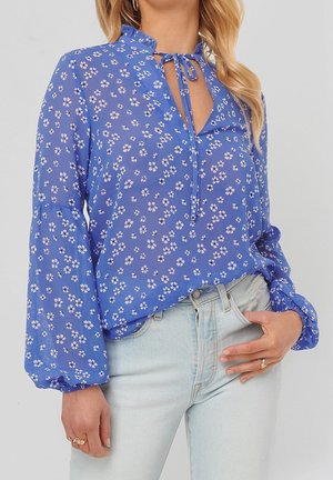 Femme portant un chemisier bleu à fleurs à manches longues avec un col à nouer et un jean délavé clair, avec des bagues aux deux mains.