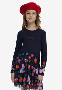 Marineblaues Langarmkleid mit tailliertem Oberteil und weit ausgestelltem Rock; farbenfrohe Früchte- und Schmetterlingsdrucke; gesticktes Logo auf der Brust. Rotes Barett.