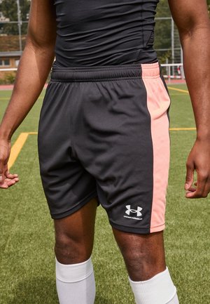 Sportowiec w czarno-różowych spodenkach Under Armour i białych wysokich skarpetach stojący na boisku ze sztuczną zieloną trawą.