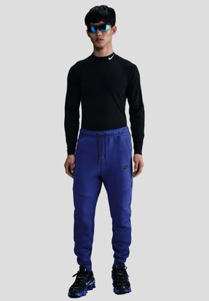 Camiseta negra de manga larga, pantalones de chándal morados con bolsillos laterales y cordón, y zapatillas deportivas negras con acentos azules. Gafas de sol con tintado azul.