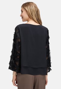 Blusa nera con maniche trasparenti e piumate; corpo liscio e scollatura arrotondata; design a strati che crea profondità e texture.