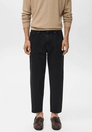 Man stands wearing a beige sweater and black jeans with cropped legs, paired with brown boat shoes, against a light gray background.