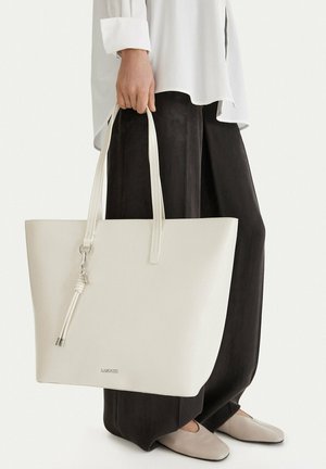 Persona che tiene una grande borsa tote bianca in pelle con manici lunghi e nappina, indossando pantaloni neri a gamba larga e top bianco.