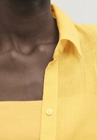 Personne à la peau foncée portant une chemise boutonnée jaune vif par-dessus un haut jaune assorti, laissant apparaître le cou et la partie supérieure de la poitrine.