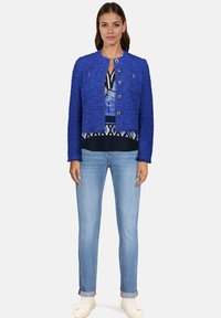 Veste texturée bleu vif au design court, avec un col rond et deux boutons à l'avant, associée à un top imprimé à plusieurs couches et un jean.