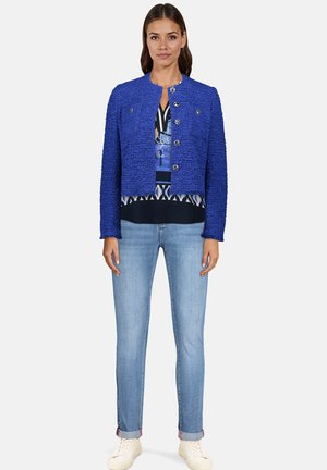 Veste texturée bleu vif au design court, avec un col rond et deux boutons à l'avant, associée à un top imprimé à plusieurs couches et un jean.