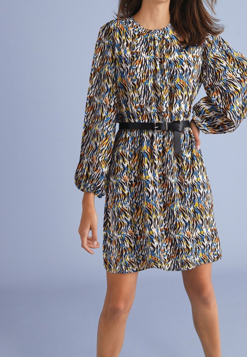 Femme portant une robe multicolore à imprimé animalier avec de longues manches bouffantes et une ceinture noire, debout devant un fond bleu clair uni.