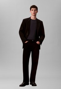 Schwarzer Blazer über einem marineblauen Hemd, kombiniert mit schwarzer Hose und Schuhen. Der Stoff ist glatt, und der Schnitt ist maßgeschneidert mit einer strukturierten Silhouette.