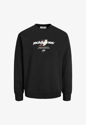 Jeune homme portant un sweat-shirt noir avec logo, pantalon olive, bottes beige et casquette noire, debout devant un fond clair uni, souriant légèrement.