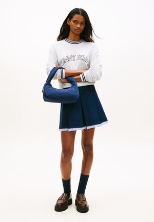 Joven de pie con los brazos cruzados, vistiendo una sudadera blanca "Tommy Jeans", falda plisada azul marino, calcetines negros, mocasines y sosteniendo un bolso azul.