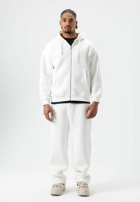Hoodie blanc à fermeture éclair avec cordons, doté d'une poche avant. Associé à un pantalon de survêtement blanc assorti et des chaussures beiges. Texture lisse.