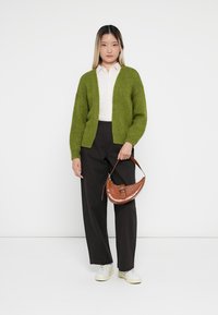 Frau steht da und trägt eine grüne Strickjacke, ein weißes Shirt, schwarze weit geschnittene Hosen und weiße Sneakers, während sie eine braune halbmondförmige Handtasche an ihrer Seite hält.