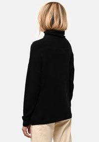 Schwarzer Rollkragenpullover, lässige Passform, weicher Stoff, überschnittene Schultern, gerippte Bündchen und Rückennahtdetails, kombiniert mit hellen Hosen.