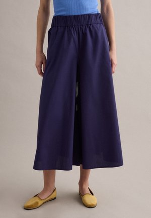 Persona con pantalones cortos de pierna ancha azul marino con cinturilla elástica, camiseta sin mangas azul brillante y zapatos tipo mocasín amarillos mostaza.