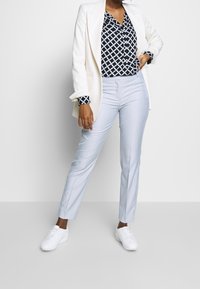 Pantalons ajustés bleu clair, associés à un chemisier à motifs noir et blanc, et une veste blanche. Des baskets blanches complètent la tenue.