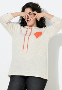 Felpa con cappuccio beige chiaro con una texture morbida, caratterizzata da un disegno a cuore arancione e coulisse. Abbinata a jeans scuri.