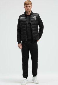 Gilet imbottito nero con colletto alto e chiusura con zip, caratterizzato da testo stampato bianco. Indossato sopra una maglietta nera a maniche lunghe e pantaloni coordinati.