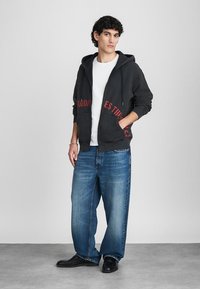 Zwart hoodie met rode letters, witte t-shirt eronder, gecombineerd met oversized blauwe spijkerbroek en zwarte schoenen, staande tegen een neutrale achtergrond.