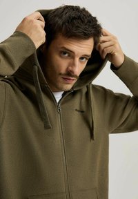 Oliwkowa rozpinana bluza z kapturem, z regulowanym sznurkiem i przednimi kieszeniami. Wykonana z teksturowanej tkaniny, z małym logo na klatce piersiowej.
