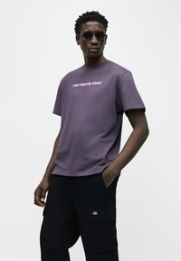 Mann trägt runde Sonnenbrille, ein lila "The North Face" T-Shirt und schwarze Cargo-Hosen, mit den Händen in den Taschen, steht vor einem schlichten Hintergrund.