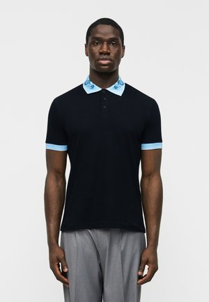 Férfi áll előre nézve, fekete polót visel világoskék gallérral és ujjvégekkel, szürke redős nadrággal párosítva.